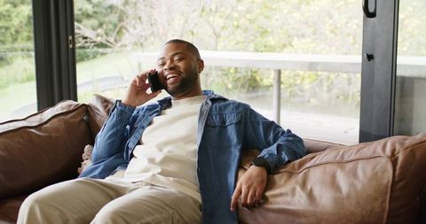 African american man reclining on leather sofa talking on smartphone and smiling on balcony