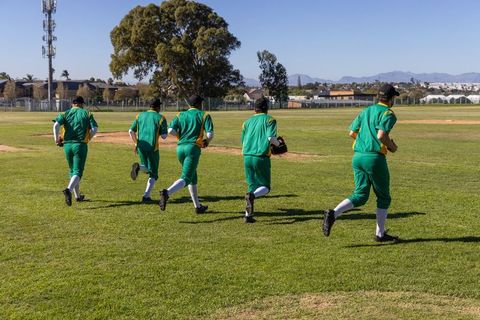 Baseball team jogging warmup drill in green uniforms across sunny outfield toward infield
