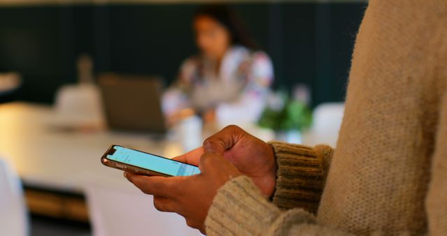 Person holding a smartphone, texting or browsing in a cozy workspace with blurred background. Ideal for illustrating modern communication, business environments, remote work, technology in daily life, or casual office settings.