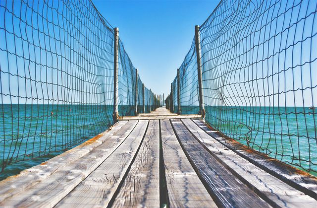Wooden pier-leading to horizon surrounded by net fencing over calm ocean waters with clear blue sky. Ideal for use in travel brochures, coastal lifestyle blogs, indoor decorations, and marine-themed websites.
