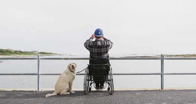 Caucasian man wearing cap sitting in wheelchair taking photo of view with dog at seaside. Disability, animals, nature and vacation, unaltered.