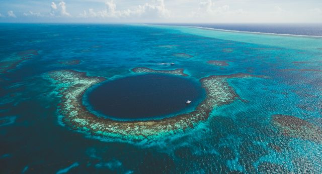 Aerial view of the Great Blue Hole in Belize. This mesmerizing marine sinkhole is surrounded by a vibrant coral reef in turquoise waters. Suitable for travel guides, ecological studies, marine exploration features, vacation promotions, and environment-related articles.