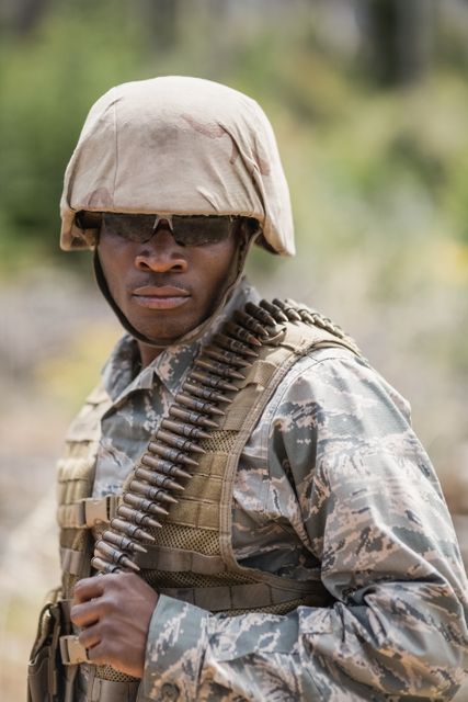 Military soldier standing outdoors in boot camp, wearing camouflage uniform and helmet, with ammunition draped over shoulder. Ideal for use in articles about military training, defense forces, and tactical gear. Suitable for educational materials, recruitment campaigns, and military-themed content.
