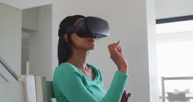 This image depicts a woman sitting indoors and using a virtual reality headset. Her finger suggests she is interacting with the VR environment, indicating an immersive and engaging experience. This image is ideal for illustrating technological advances, virtual reality applications, digital transformation, and modern home settings involving today's cutting-edge gadgets.