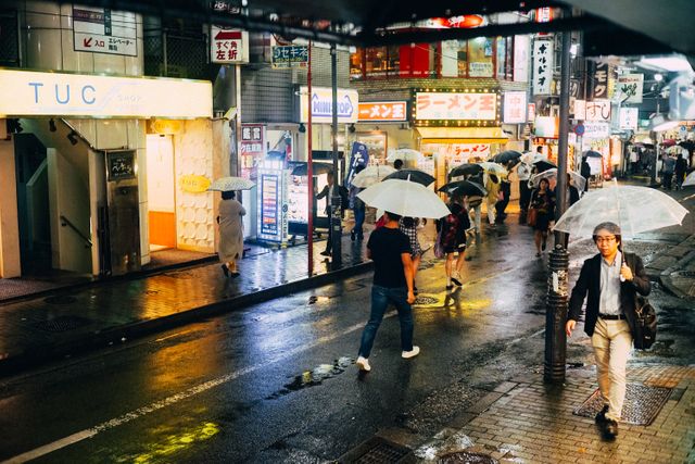 Busy Tokyo street filled with people holding umbrellas during a rainy night. Illuminated signs, wet pavement, and urban atmosphere reflect vibrant city life. Perfect for projects related to urban living, travel in Japan, nightlife, and rainy weather mood.