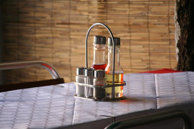A close-up of a condiment set including salt, pepper, vinegar, and oil on a dining table in a restaurant. The background features wooden blinds, adding a cozy atmosphere to the indoor setting. Suitable for use in articles or promotions related to dining, culinary tools, restaurant decor, or food services.