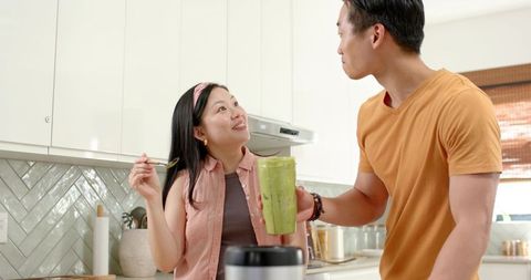 Asian couple tasting green smoothie in bright modern kitchen holding blender and spoon