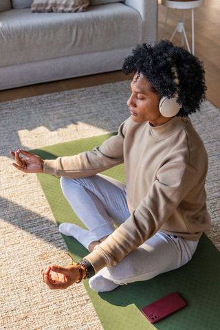 Black woman practicing meditation with headphones on green yoga mat in sunlit living room