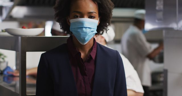 Restaurant manager stands confidently in commercial kitchen wearing a protective mask. Background chefs are blurred, focusing attention on the manager. Useful for topics related to health safety, food industry, restaurant management, workplace safety, and professional environments.