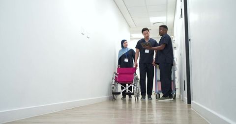Diverse nursing team reviewing tablet while guiding wheelchair in bright hospital corridor
