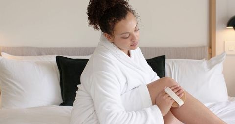 African american woman exfoliating legs in white bathrobe while sitting on bed with pillows