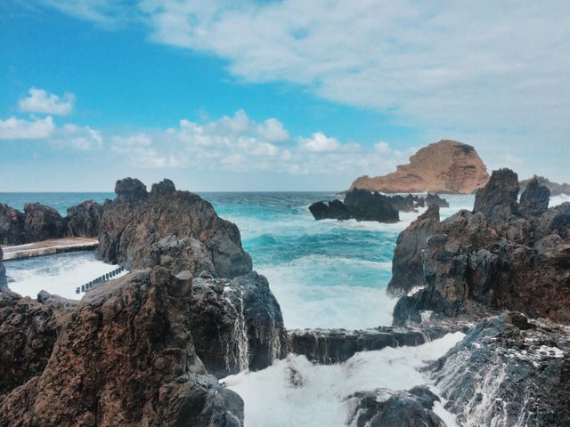 Natural rock pools surrounding by rugged rocks on a coastline under a bright blue sky with scattered clouds. Ideal for use in travel guides, nature documentaries, coastal adventure promotions, or as a serene desktop background emphasizing natural beauty and tranquility.