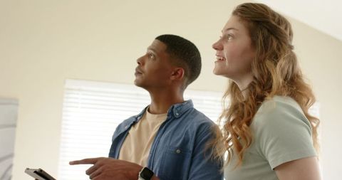 Diverse couple inspecting new home with tablet, looking up in bright modern interior space
