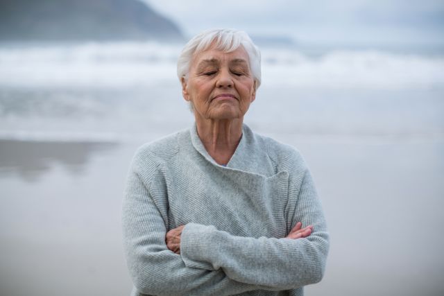 Senior woman standing on beach with eyes closed, arms crossed, meditating. Ideal for promoting mental health, wellness, mindfulness, and relaxation. Suitable for use in articles, blogs, and advertisements focused on elderly care, self-care, and peaceful living.