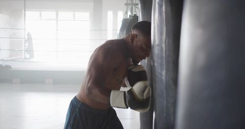 Shirtless boxer leaning on heavy bag during intense training session in sunlit gym