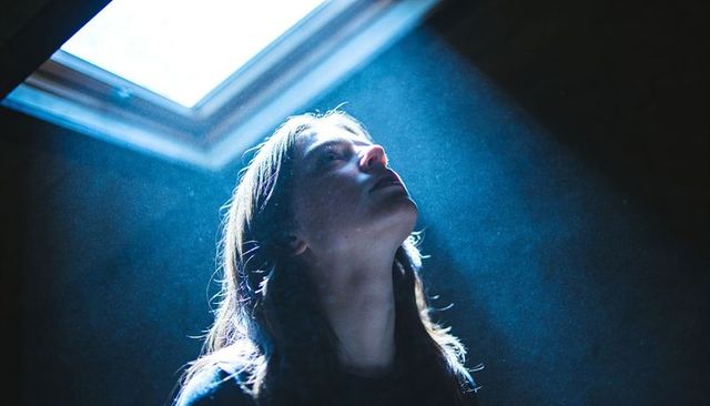 Woman gazing upward into skylight beam in moody loft, cinematic rimlight and dust