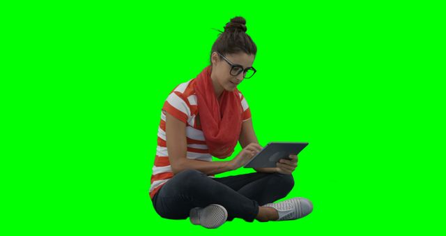 Young woman with glasses sitting cross-legged, using a digital tablet, isolated on green screen background. Suitable for technology, education, lifestyle, and marketing projects. Green screen allows for easy background replacement.
