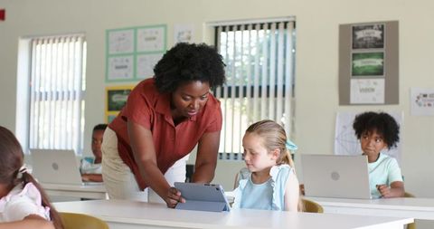 Black teacher guiding young Caucasian girl using tablet in bright classroom for learning