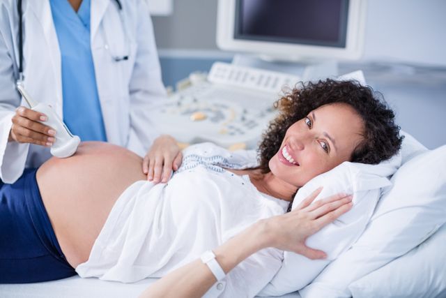 This image depicts a pregnant woman receiving an ultrasound scan in a hospital setting. The woman is smiling and appears relaxed while a healthcare professional conducts the scan. This image can be used for articles or advertisements related to prenatal care, pregnancy, maternity services, healthcare, and medical examinations.