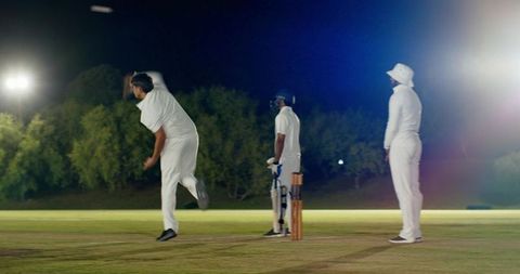 Night cricket match under floodlights, bowler delivering to batsman near stumps, diverse players