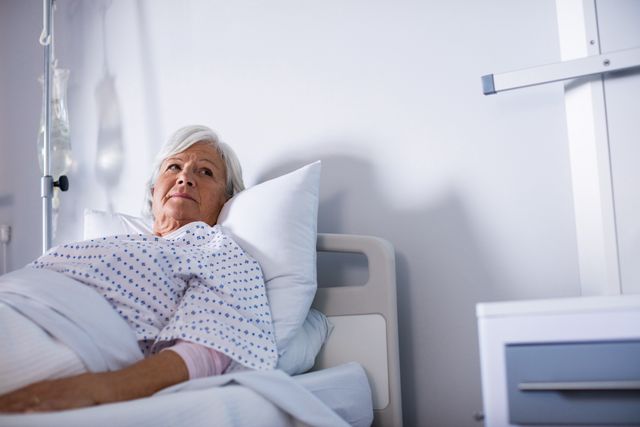 Senior woman lying in a hospital bed, looking thoughtful. She is wearing a hospital gown and has an IV drip attached. The setting is a hospital room, indicating a medical environment. This image can be used for healthcare, medical care, elderly care, recovery, and hospital-related themes.