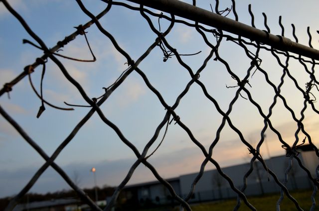 Weathered chain link fence with barbed wire seen at sunset, against background of industrial setting. Can be used in topics on urban decay, security, infrastructure, and industrial themes. Suitable for editorials, blogs, and articles discussing security, boundary issues, and urban development.