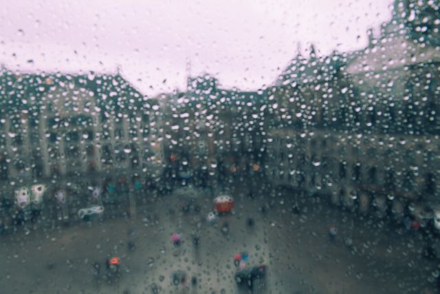 View through window glass covered with raindrops overlooking an urban square. The raindrops on the glass in focus, while the urban background is blurred. Ideal for themes of weather, rain, city life, mood, and atmospheric scenes.