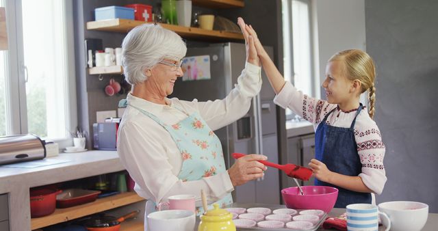 Grandmother and granddaughter enjoying quality time while baking in kitchen. They share a high five, celebrating their efforts. Suited for themes of family bonding, intergenerational relationships, cooking, and joyful moments.