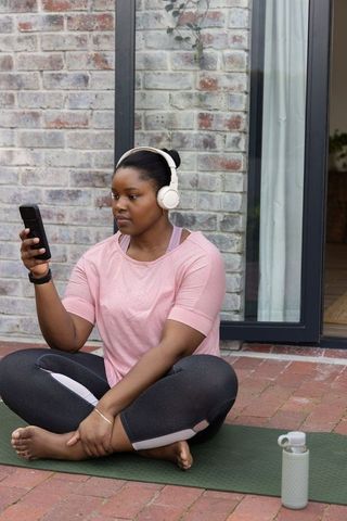 African american woman listening to music and checking smartphone on outdoor yoga mat