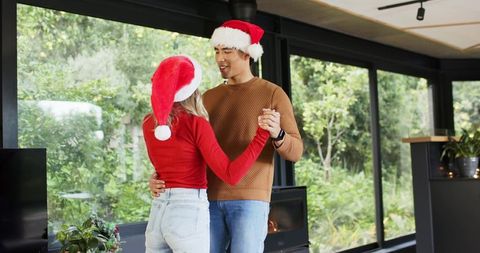 Couple dancing in cozy modern living room wearing santa hats and festive sweaters