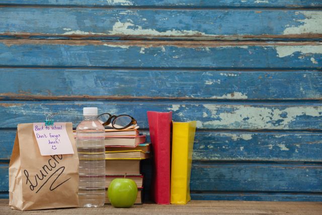 Stack of books, apple, water bottle, spectacles and lunch bag against blue wooden background