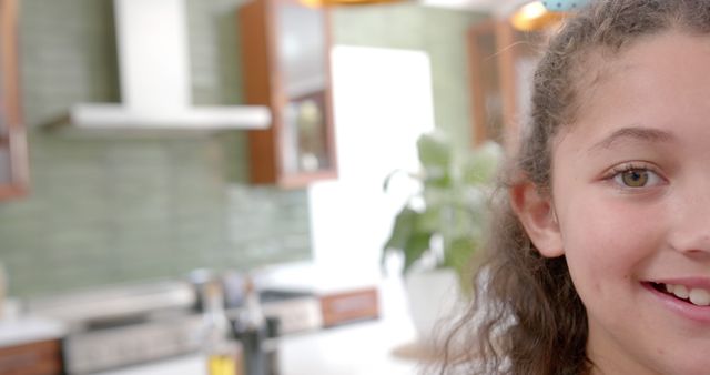 Half face of happy biracial girl with curly hair standing and smiling in sunny kitchen, copy space. Childhood, wellbeing, free time and domestic life, unaltered.