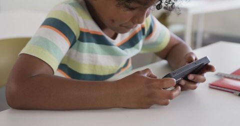 Young student playing handheld game at classroom desk with notebook and pencil