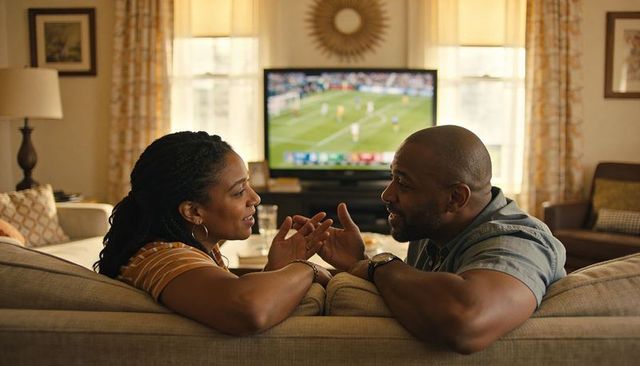 Black couple leaning on sofa talking while watching football in living room