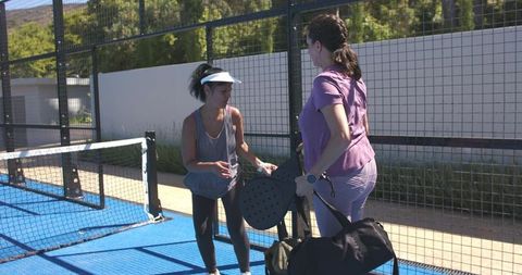 Exchanging padel paddles on sunlit blue court, women teammates preparing match and packing gear
