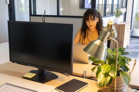 Young professional working on laptop in modern open-plan office with monitor and greenery