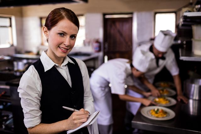 Female waitress taking an order on a notepad in a busy hotel kitchen while chefs prepare dishes in the background. Ideal for use in articles or advertisements related to the hospitality industry, restaurant management, culinary arts, and teamwork in professional kitchens.