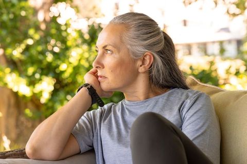 Pensive mature woman relaxing on sunlit patio sofa, looking toward lush garden
