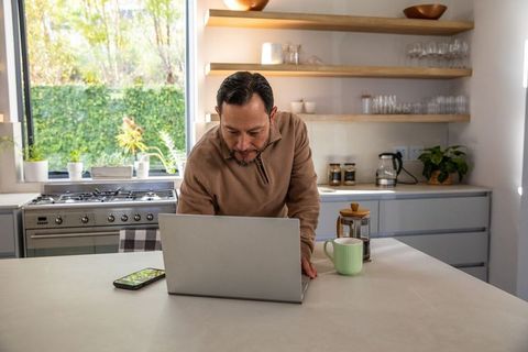 Asian man leaning on laptop in bright kitchen, working remotely with coffee and smartphone