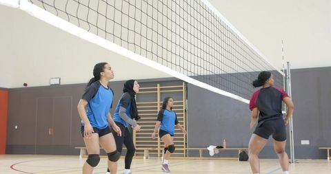 Multicultural female volleyball squad practicing under net in school gym wearing hijab, knee pads