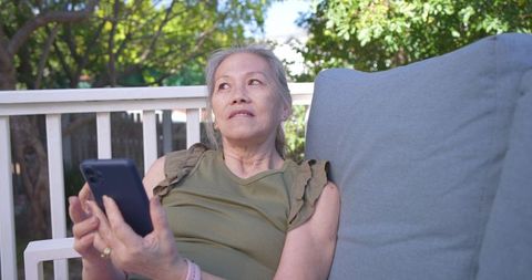 Senior asian woman relaxing on backyard deck holding smartphone, gazing upward and reflecting