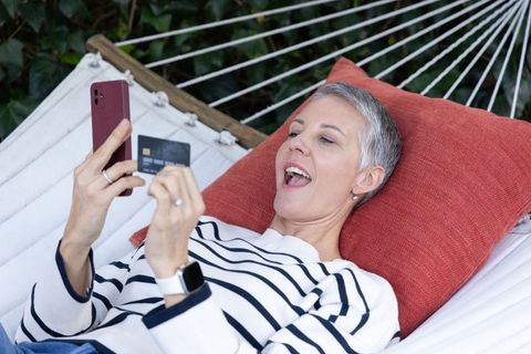 Senior woman reclining in hammock using smartphone and credit card for online shopping