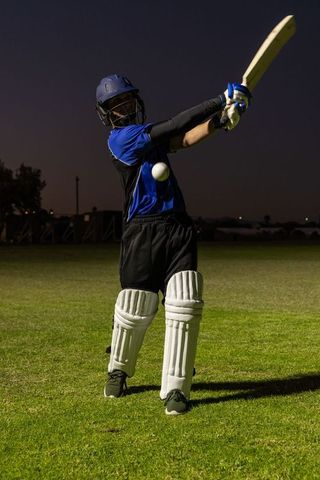Cricket batter swinging powerful shot at dusk under stadium lights wearing pads and helmet