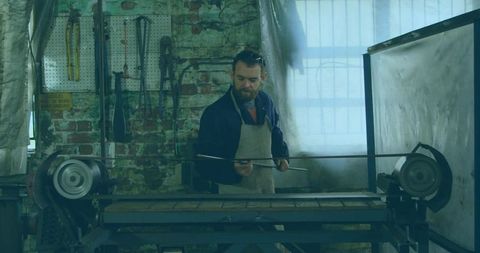 Bearded machinist feeding metal rod into industrial roller bed in vintage brick workshop