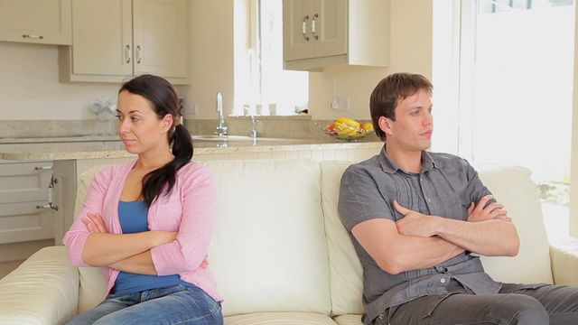 A couple with expressions illustrating unhappiness and tension sitting on a sofa with arms crossed. They are facing away from each other in what appears to be a modern living room, indicating a communication problem or relationship conflict. Useful for illustrating themes of relationship issues, domestic life struggles, and emotional complexity in personal interactions.