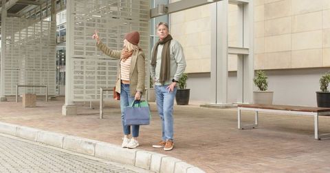 Mature couple hailing ride at curb in urban street holding tote bags wearing fall layers