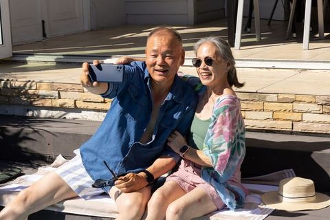 Senior asian couple taking joyful smartphone selfie on sunlit patio during summer vacation