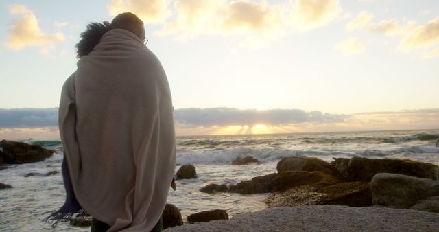 Romantic diverse couple with blankets on backs standing on beach and looking at sunrise. Summer, vacation, romance, love, relationship, free time and lifestyle, unaltered.