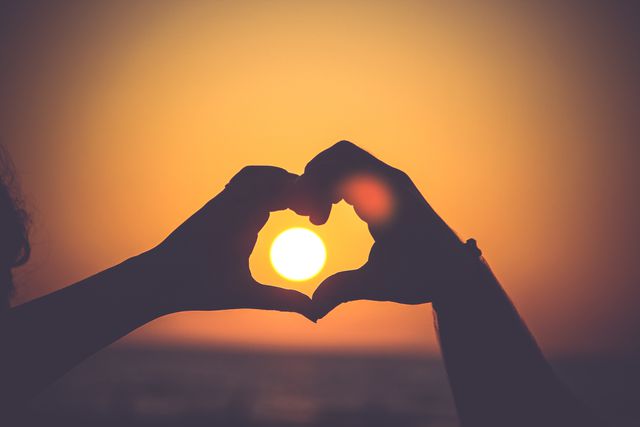 This image shows a pair of hands creating a heart shape against a stunning sunset background, with the sun peering through between the fingers. The photo's warm colors and romantic theme make it perfect for use in advertising for travel destinations, romantic getaways, Valentine's Day promotions, wedding-related publications, or any material aimed at evoking feelings of love and connection.