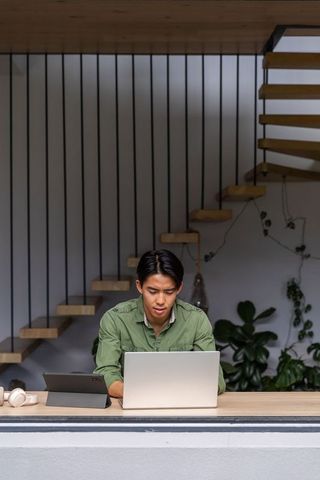 Asian man working on laptop at wooden counter in modern home office with staircase